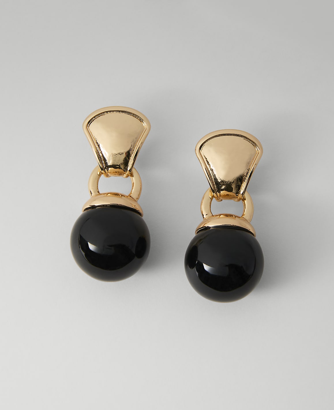 A pair of gold earrings with black pearl drops hang against a plain, light gray background.