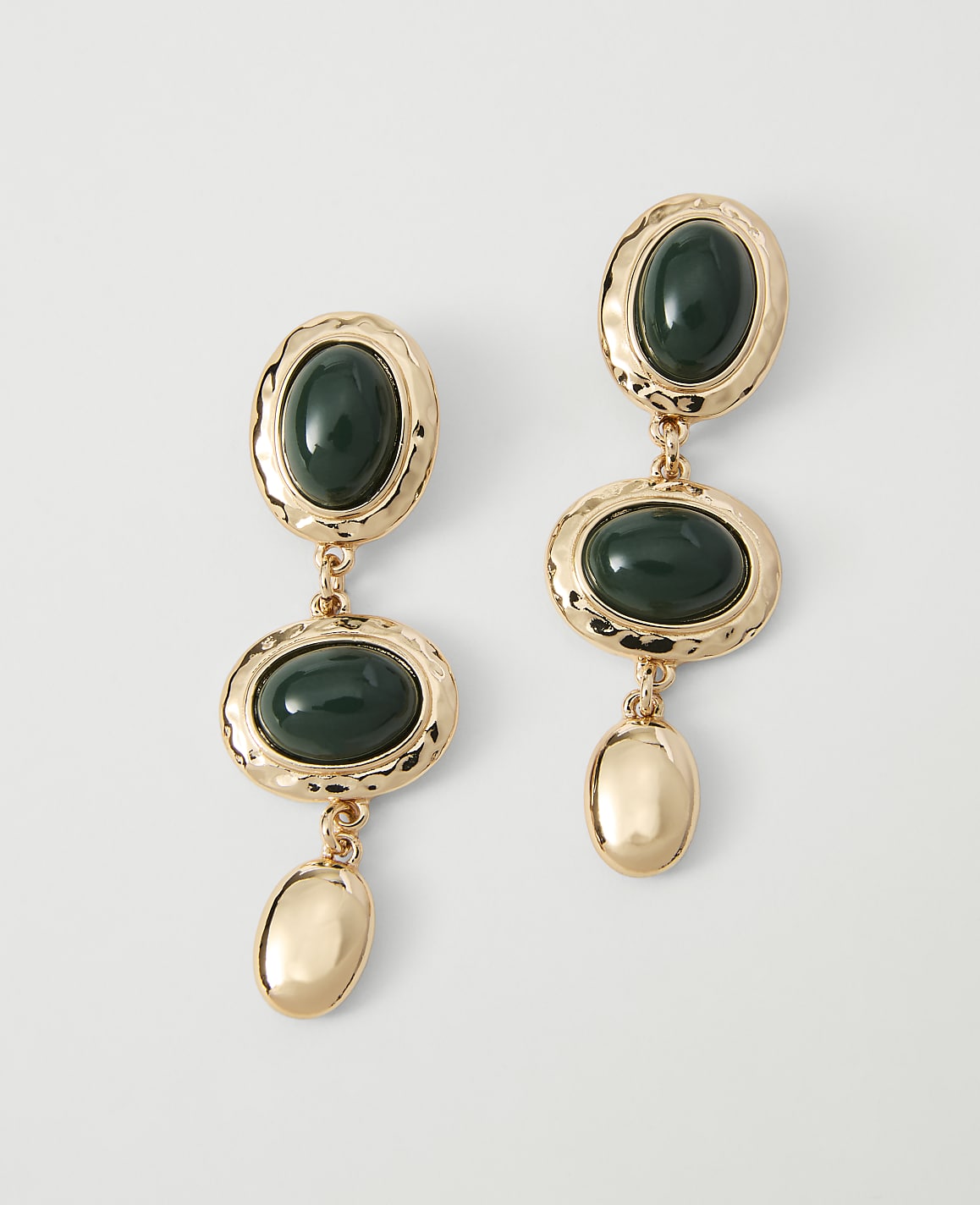 A pair of gold dangle earrings with oval green stones and gold accents hang against a plain white background.