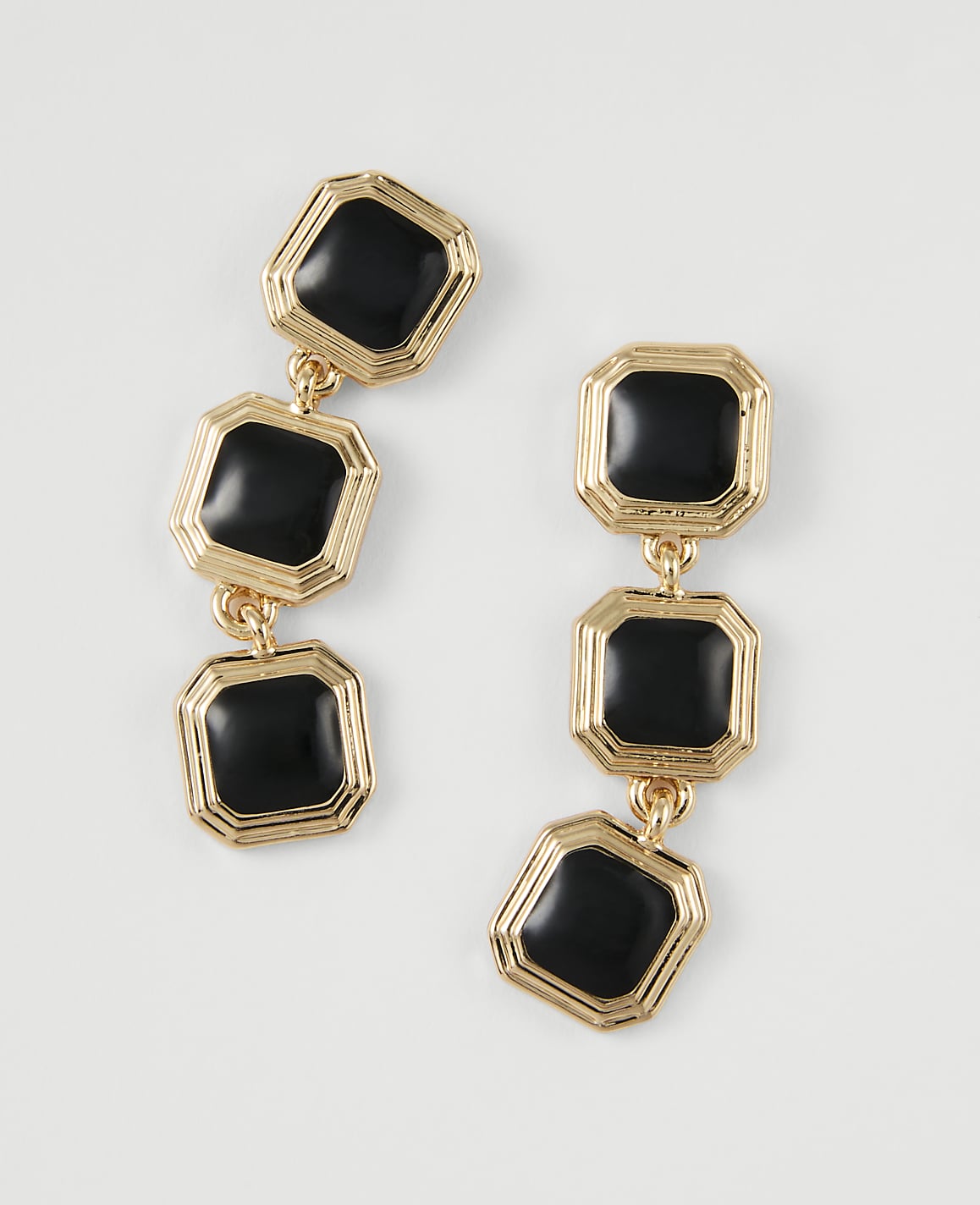 A pair of gold-toned dangle earrings with three black square charms each are laid out on a plain white background.