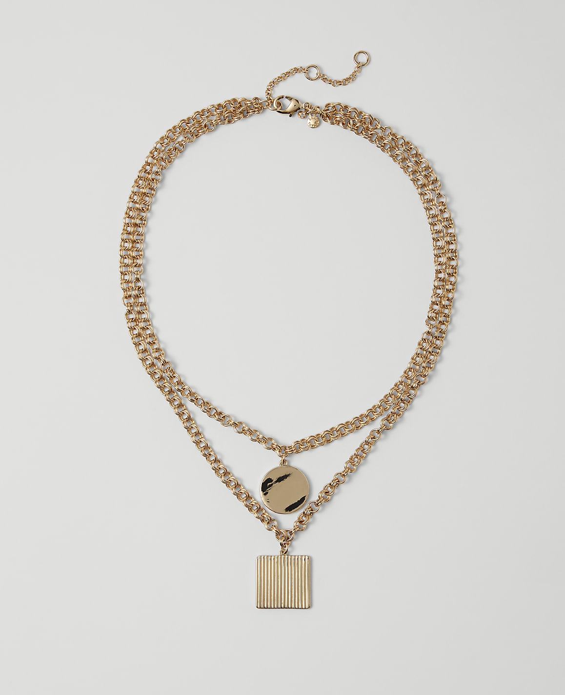 A layered gold necklace with a circular pendant and a square pendant hangs against a plain white background.