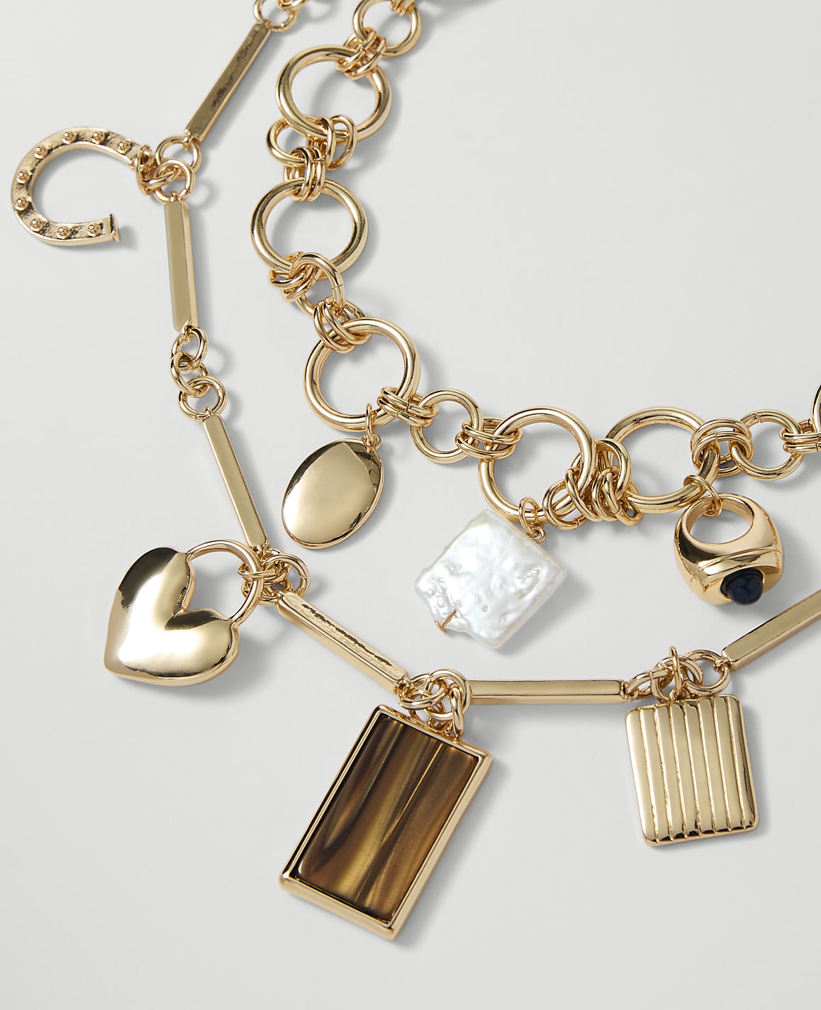 A gold necklace with various charms, including a heart, horseshoe, and rectangular pendants, rests against a plain white background.