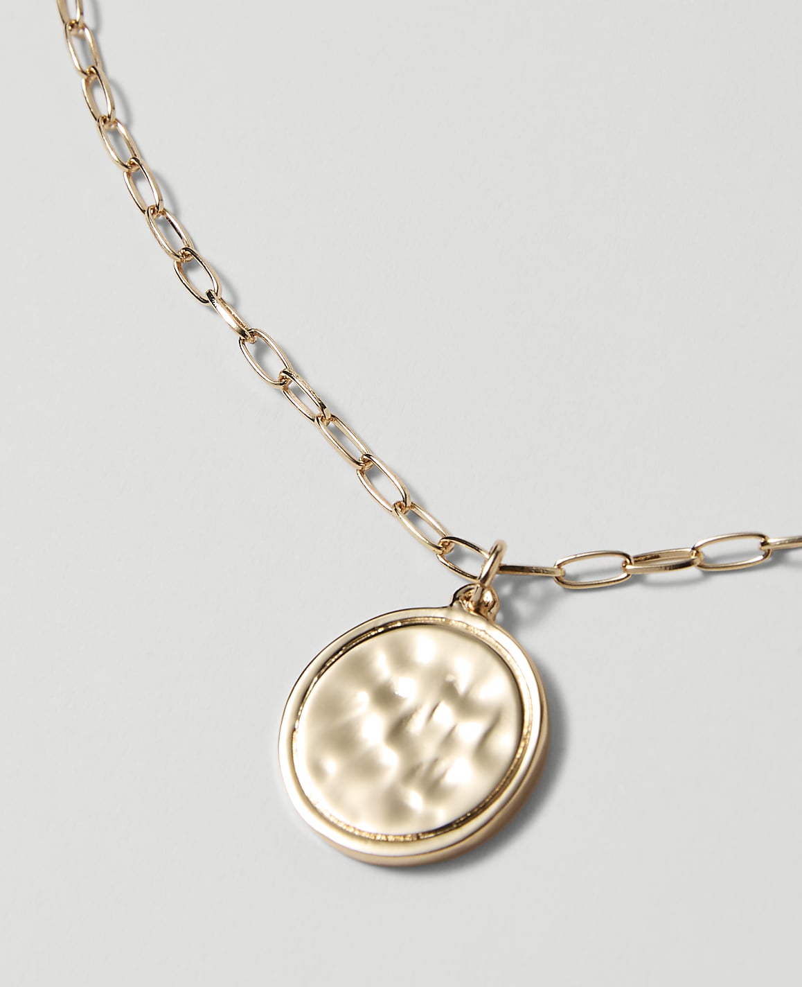 A gold hammered coin pendant hangs from a delicate gold chain against a plain, light gray background.