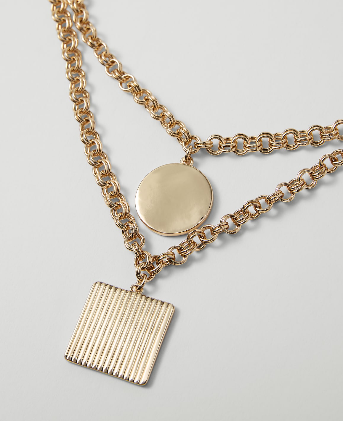 A gold necklace with a square pendant in the foreground rests against a plain, light gray background.
