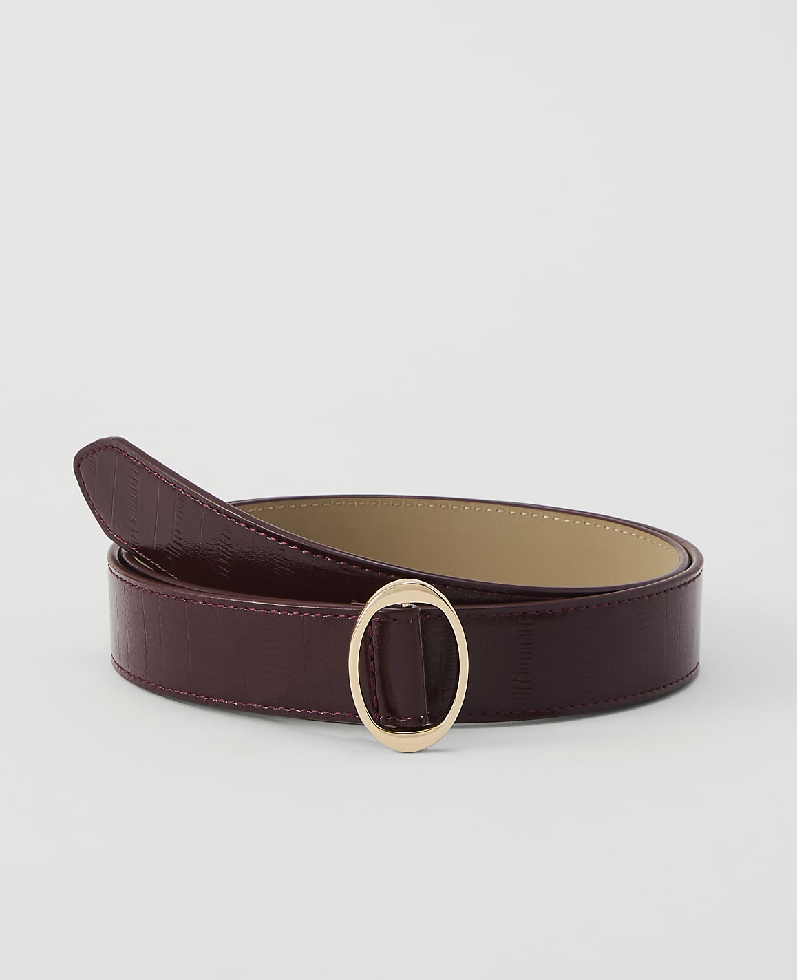 A burgundy leather belt with a gold oval buckle lies on a plain white background.