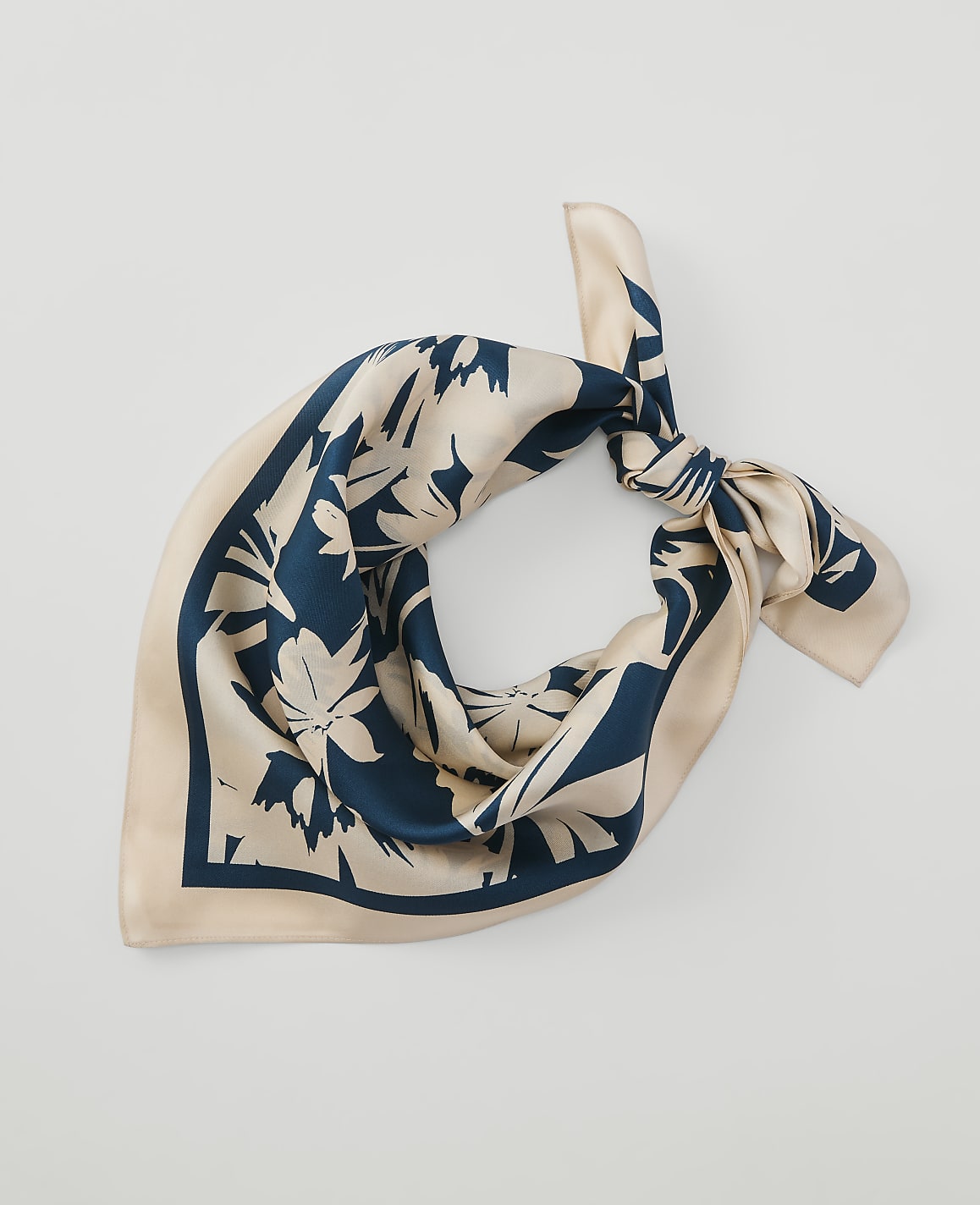 A cream-colored silk scarf with a dark teal floral pattern is tied in a knot against a plain white background.