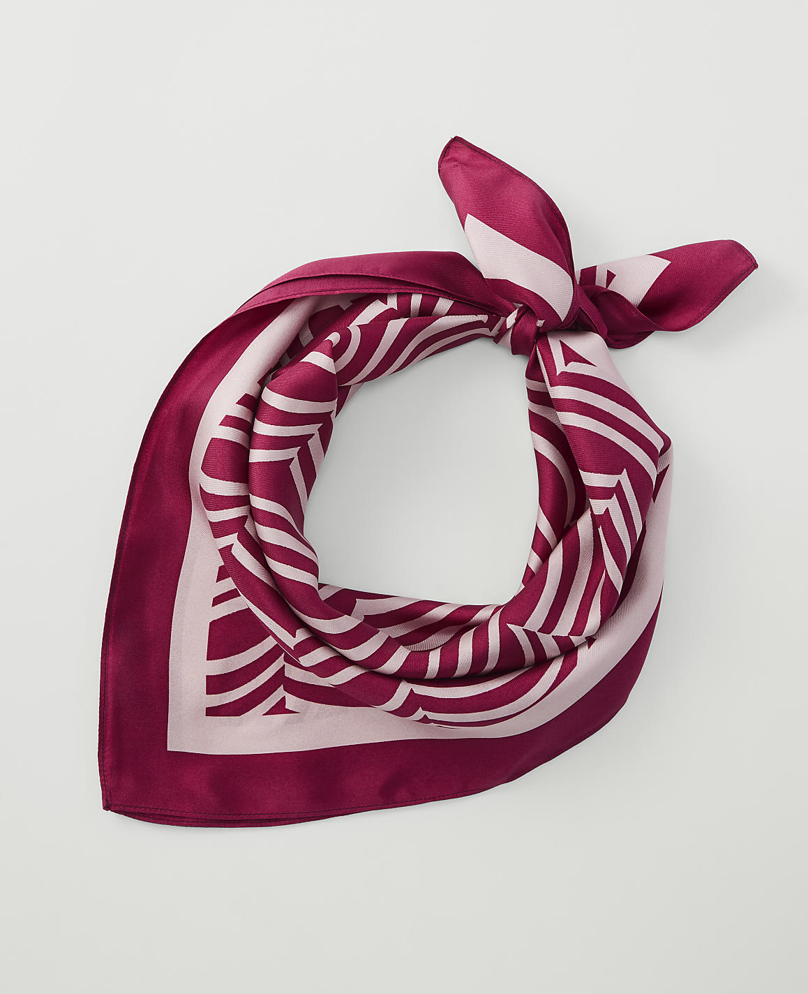 A maroon and pink patterned silk scarf is tied in a knot in the foreground against a plain white background.