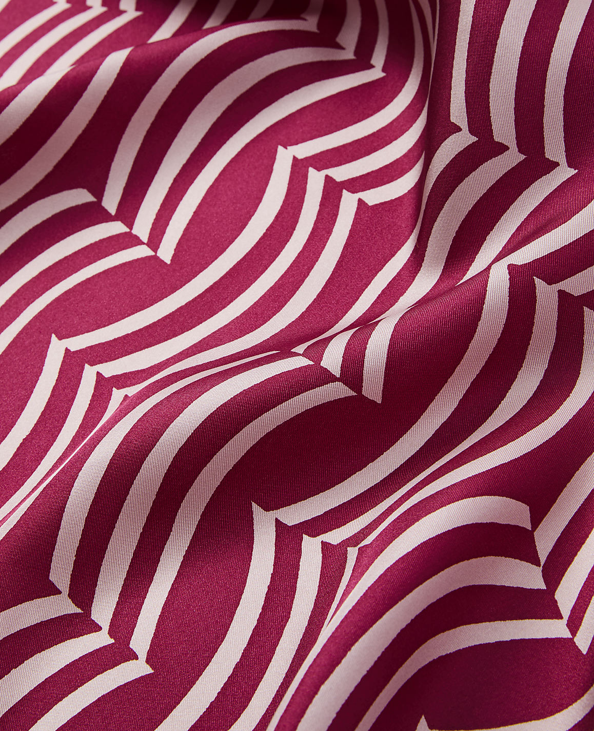 A close-up of a flowing, deep pink fabric with a repeating pattern of cream-colored, wavy lines creates a dynamic and textured foreground against a softly shadowed background.