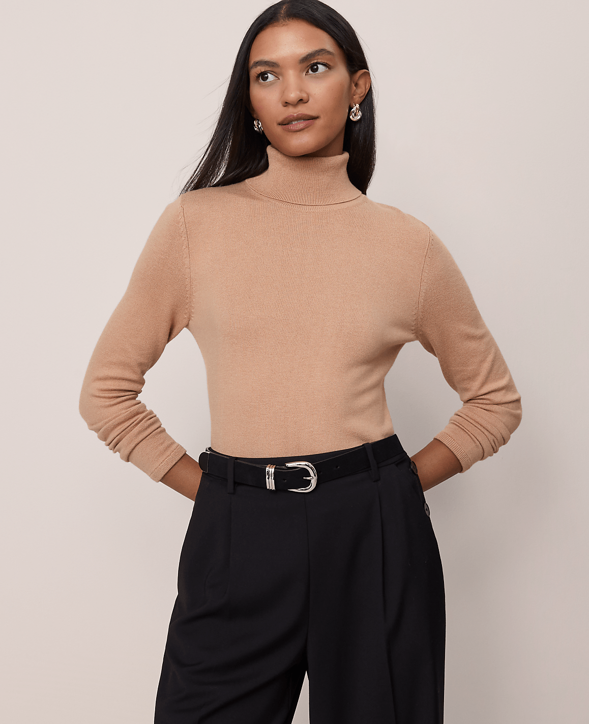 A woman in a tan turtleneck and black pants stands against a plain beige background.