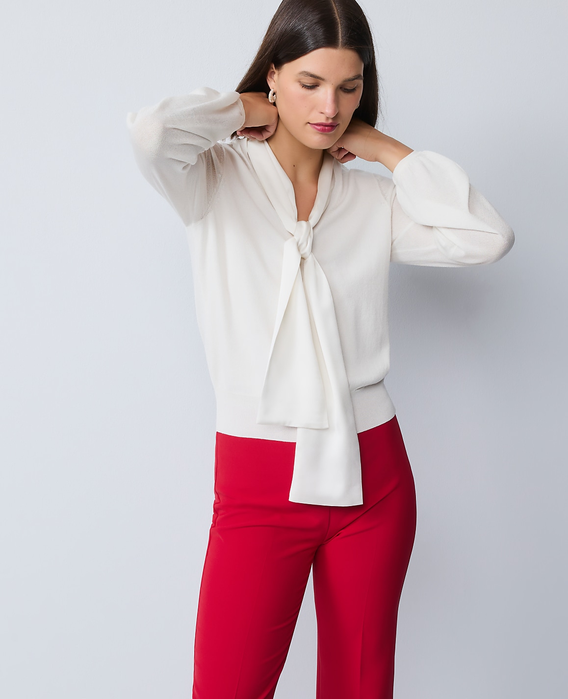 A woman in a cream bow-neck blouse and red pants stands against a plain gray background.