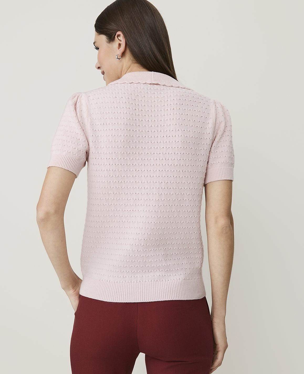 A woman in a pink collared shirt and maroon pants stands against a plain white background.
