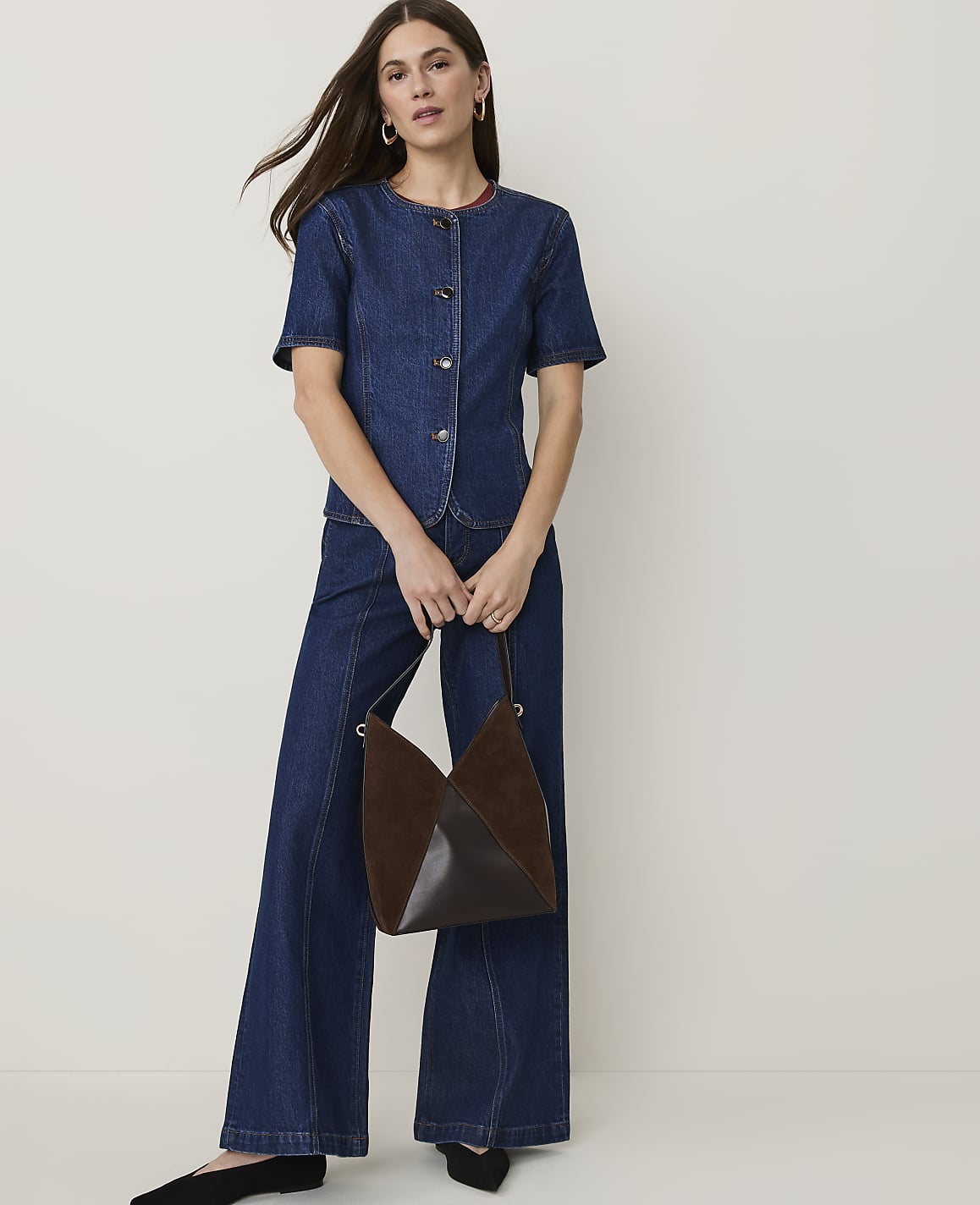 A woman in a denim outfit stands holding a brown handbag against a plain white background.