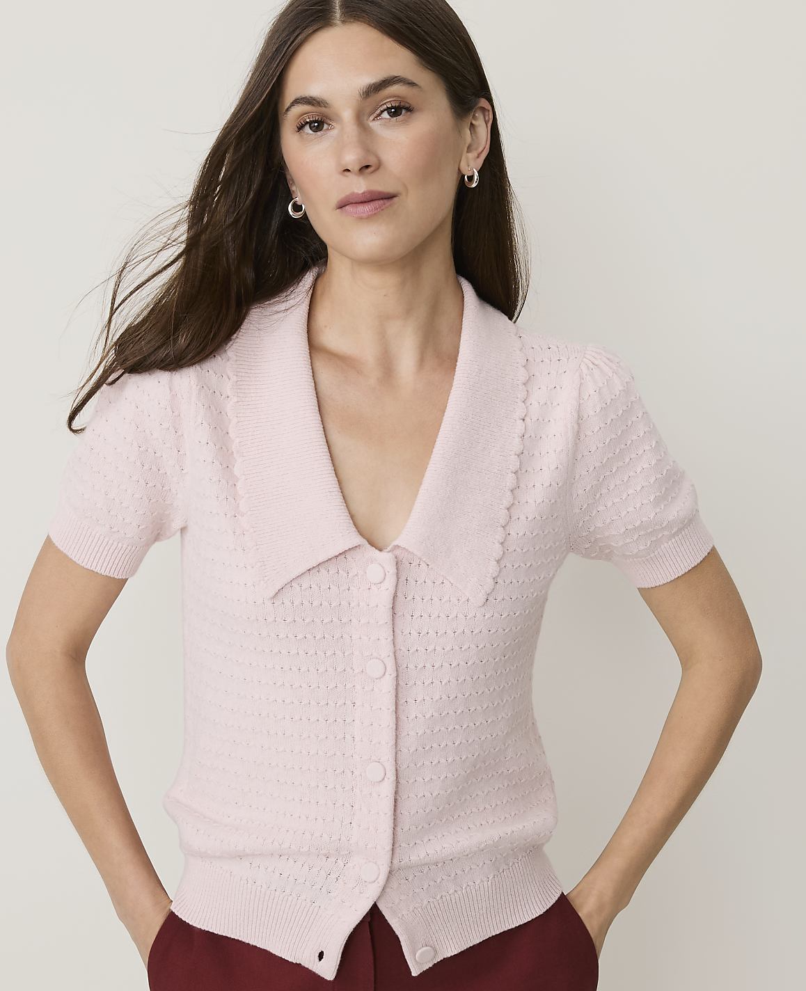 A woman in a pink collared cardigan stands against a plain white background.