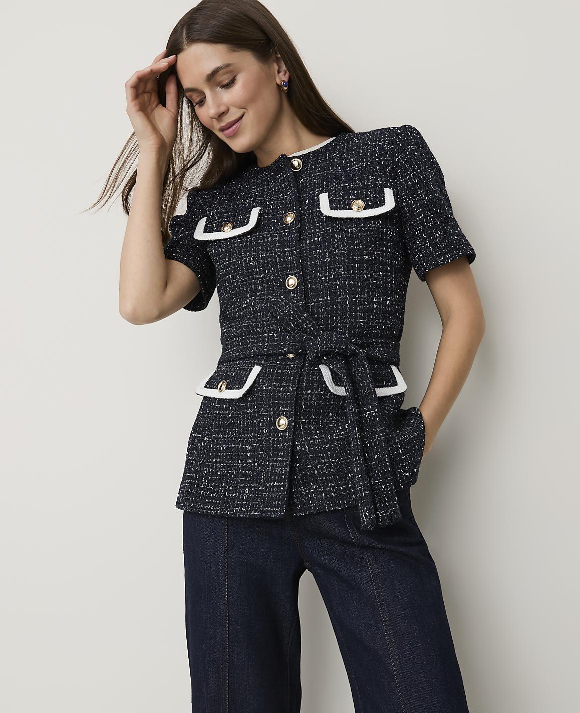 A woman in a dark tweed jacket with white trim and a belt stands against a plain, light-colored background.