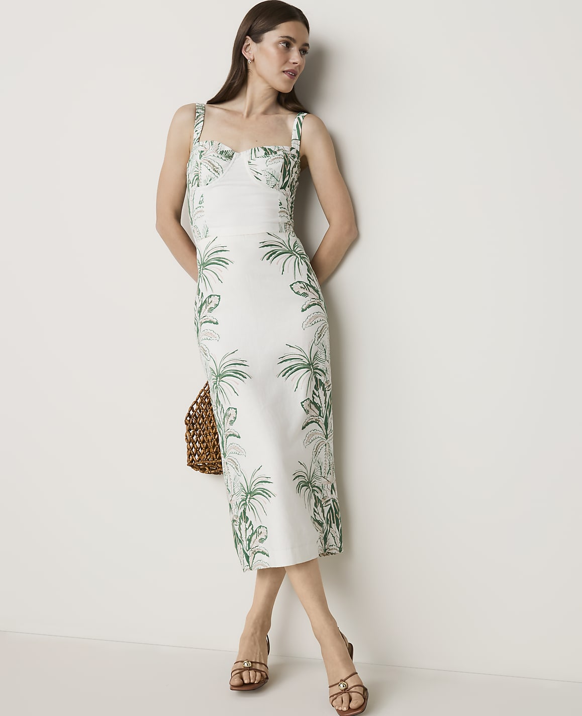 A woman in a white dress with a tropical palm print stands against a plain white wall, holding a woven bag.