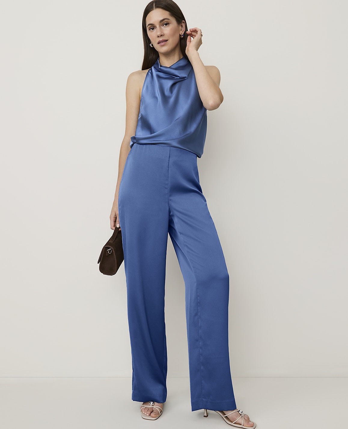 A woman in a blue satin halter top and wide-leg pants poses with a brown clutch against a plain, light background.
