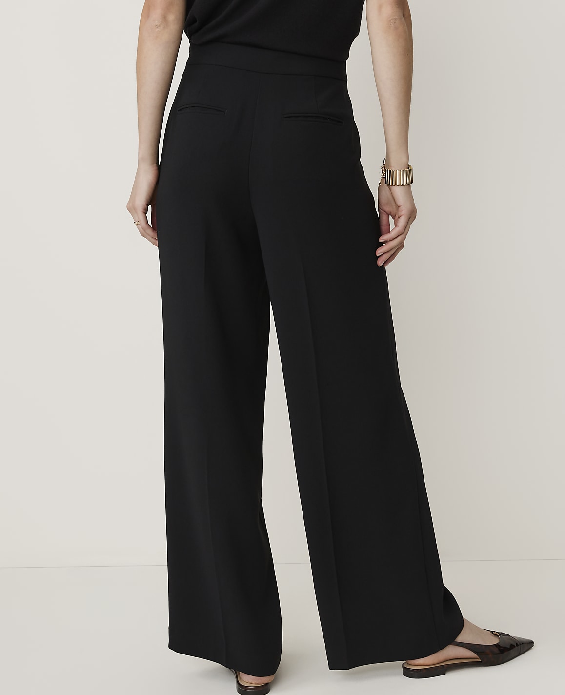 A person wearing black wide-leg pants and pointed-toe flats stands against a plain, light-colored background.