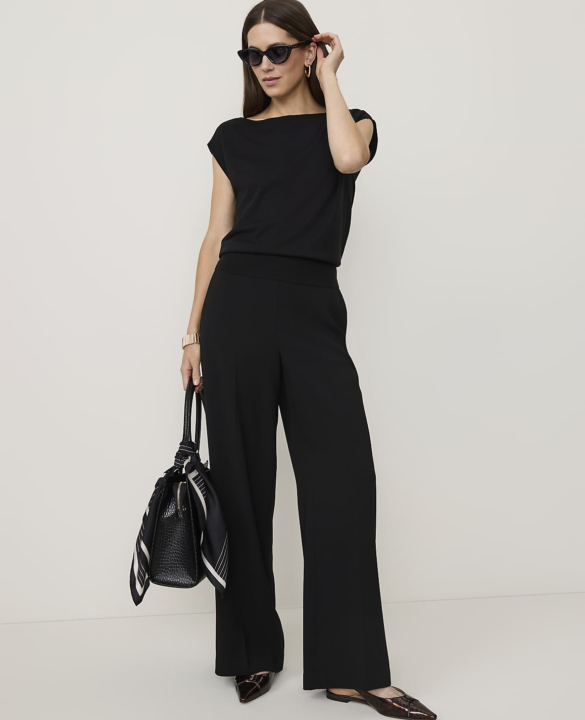 A woman in a black jumpsuit and sunglasses stands with a handbag in front of a plain white background.
