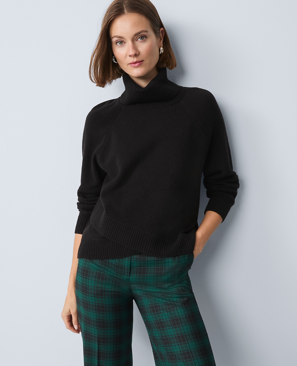 A woman in a black turtleneck sweater and green plaid pants stands against a light blue background.