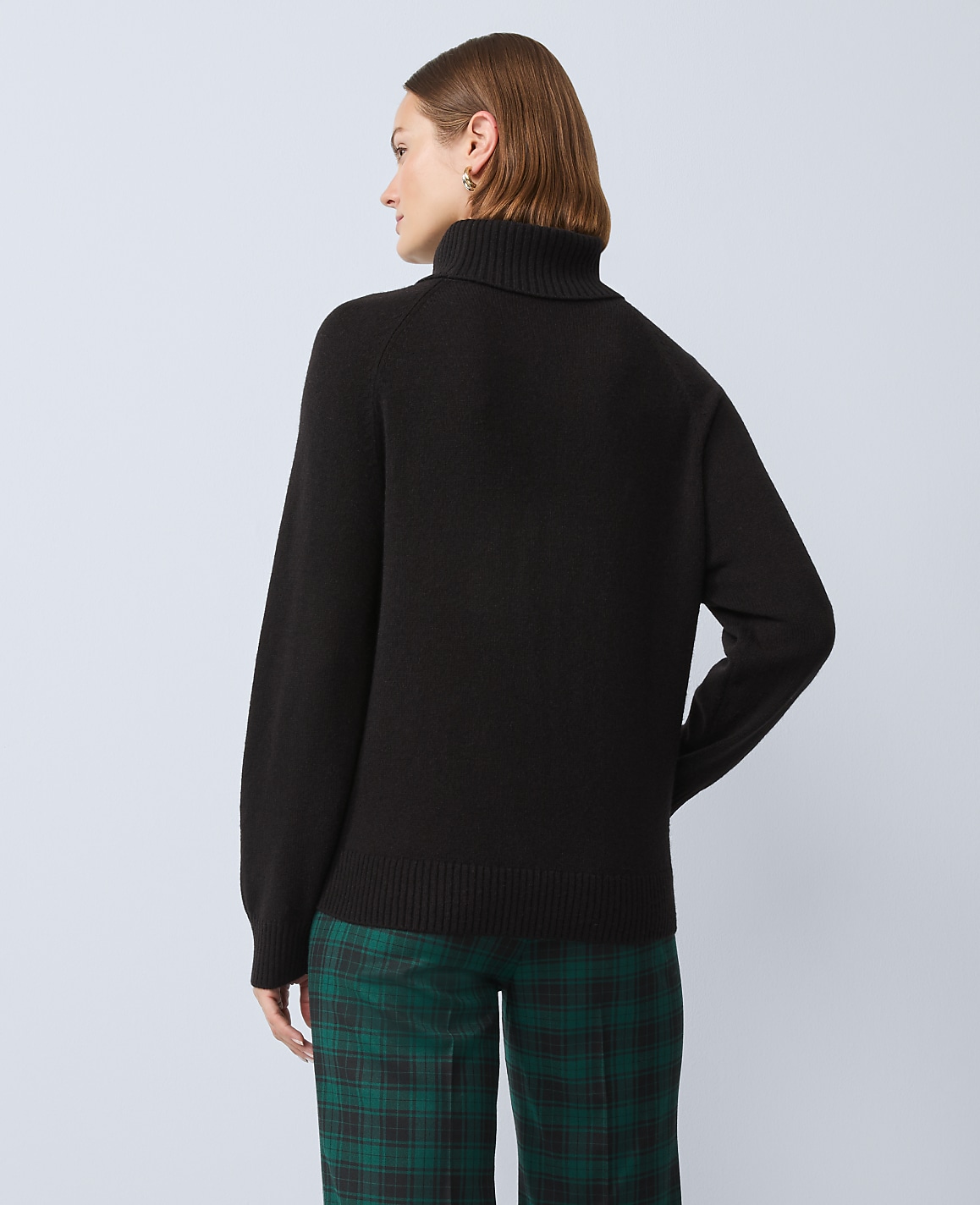 A woman in a black turtleneck sweater and green plaid pants stands against a plain gray background.