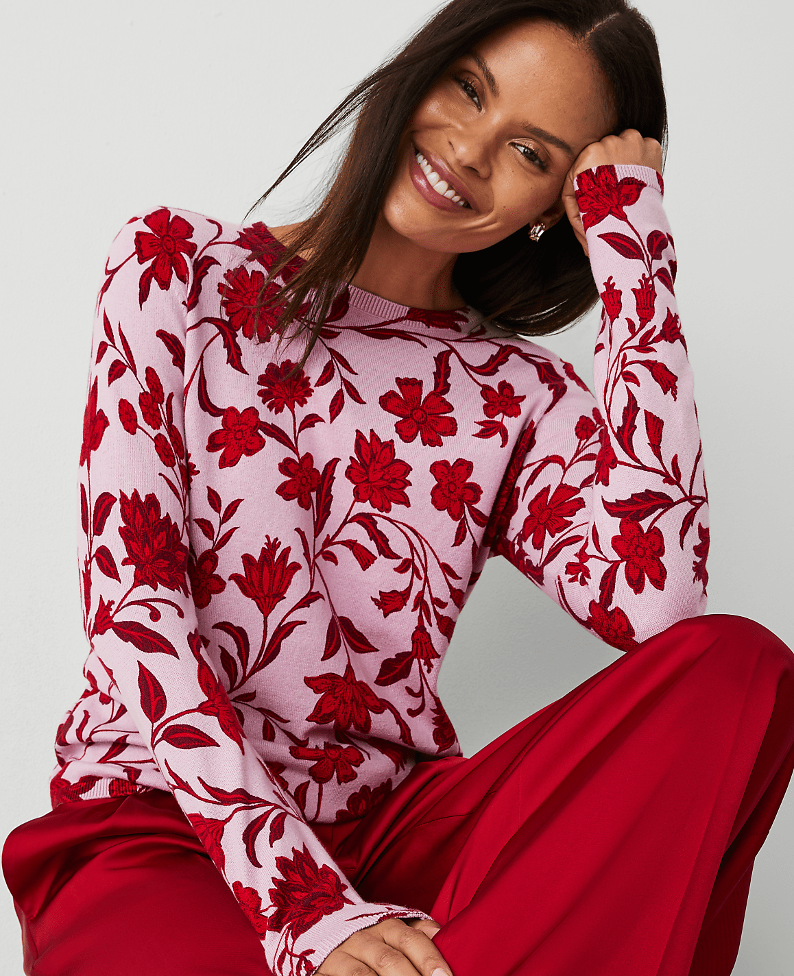 A smiling woman in a pink floral sweater and red pants sits against a plain background.