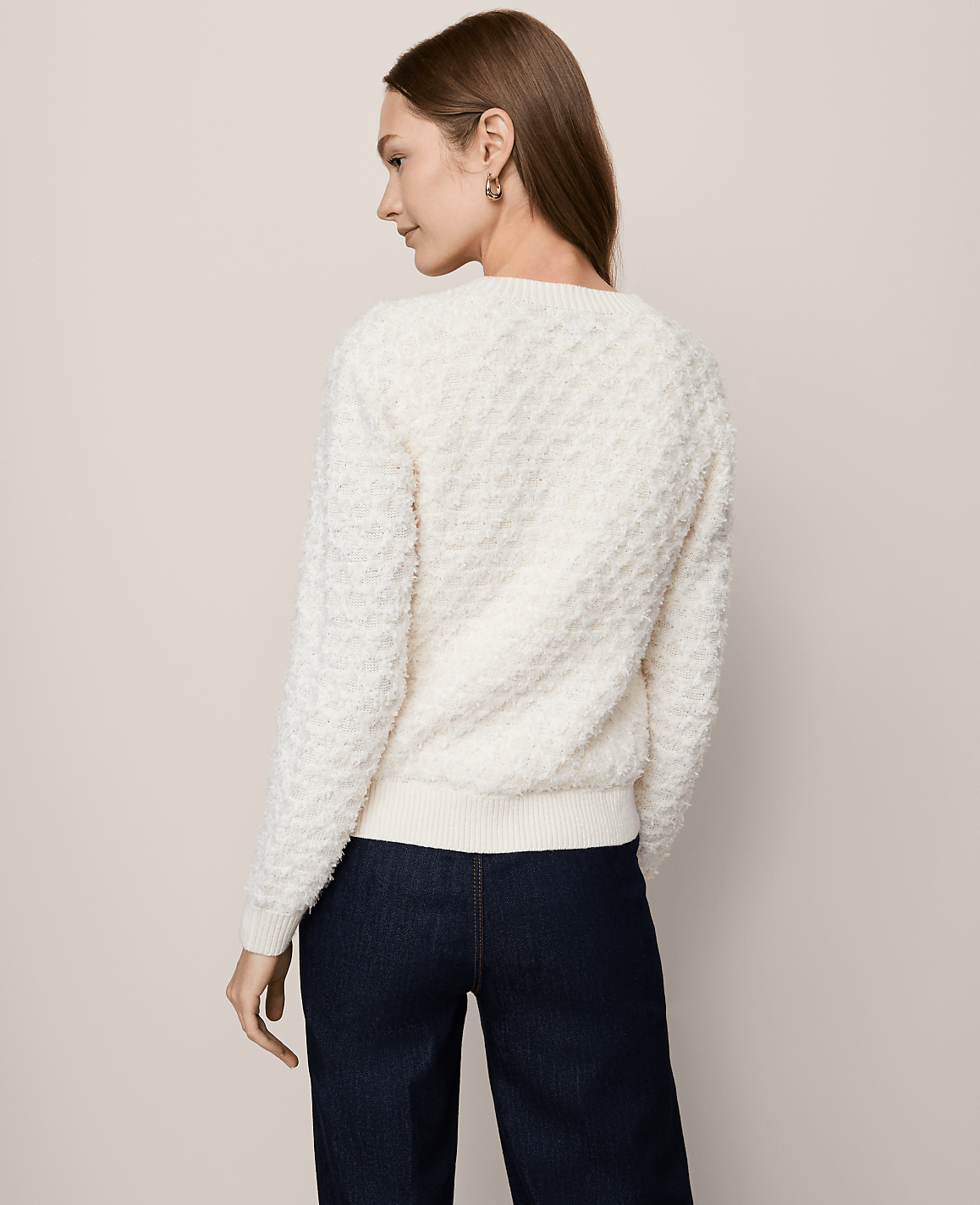 A woman in a textured cream sweater and dark jeans stands against a plain, light-colored background.