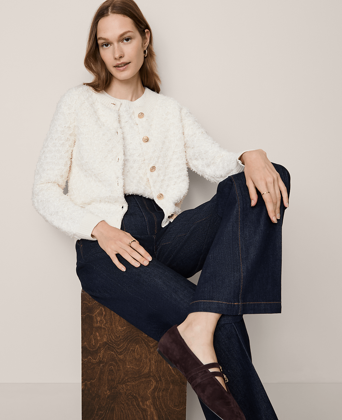 A woman in a cream textured cardigan and dark wide-leg jeans sits on a wooden stool against a plain, light background.