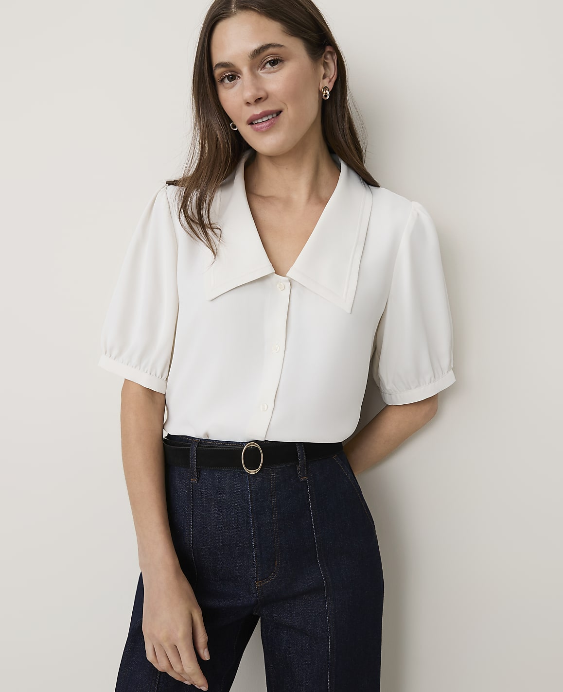 A woman in a white blouse and dark jeans stands against a plain white background.