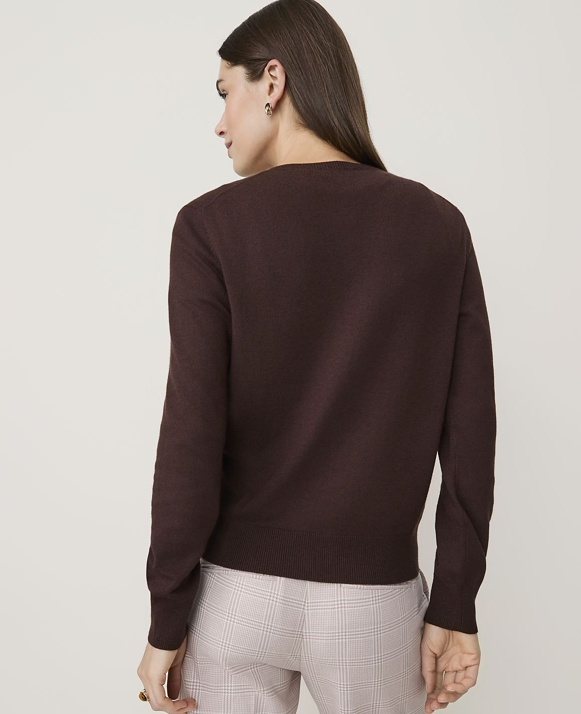 A woman in a brown sweater and plaid pants stands against a plain, light-colored background.