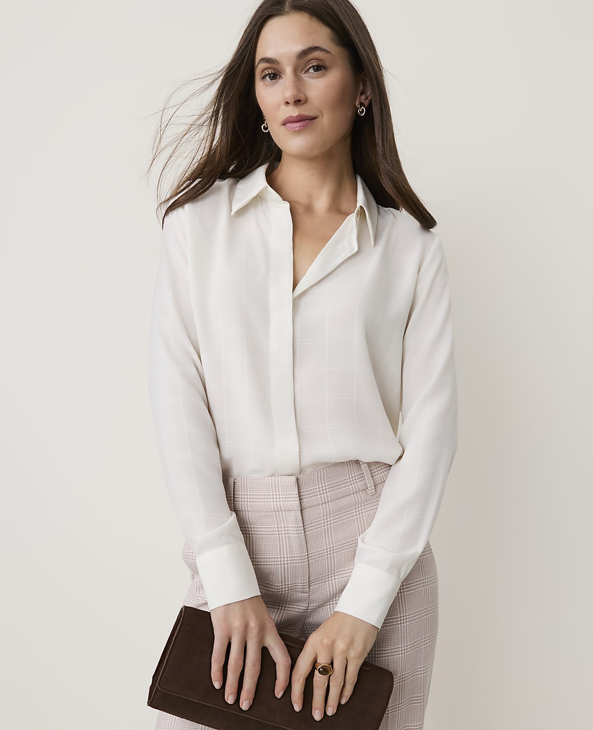 A woman in a cream blouse and plaid pants holds a dark clutch in front of a plain background.
