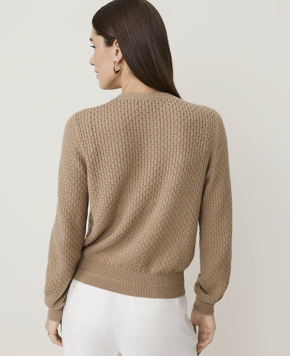 A woman in a tan textured sweater and white pants stands against a plain, light-colored background.