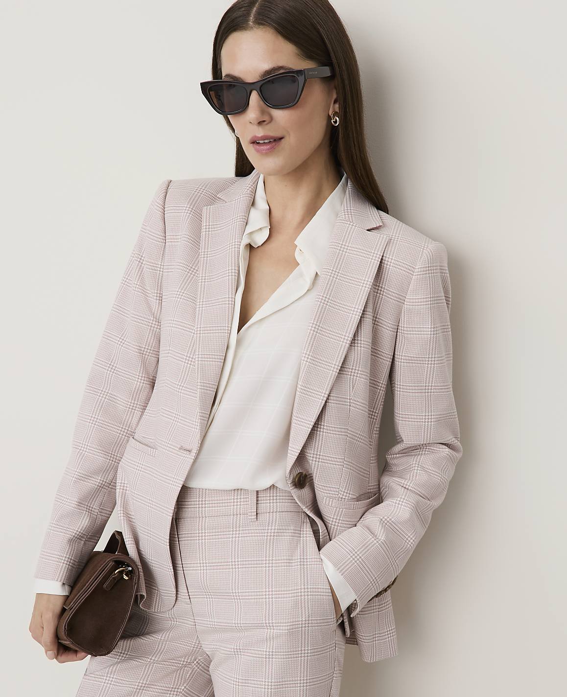 A woman in a pink plaid suit and sunglasses holds a brown clutch in front of a plain white background.