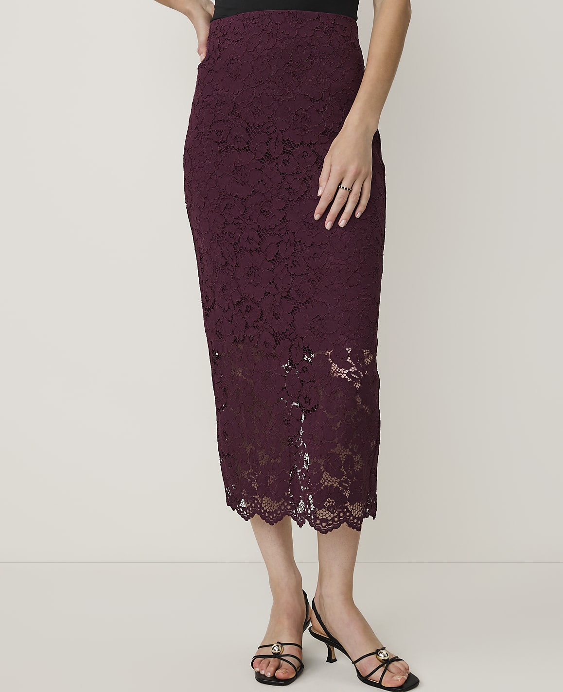 A woman in a burgundy lace pencil skirt and black heels stands against a plain white background.