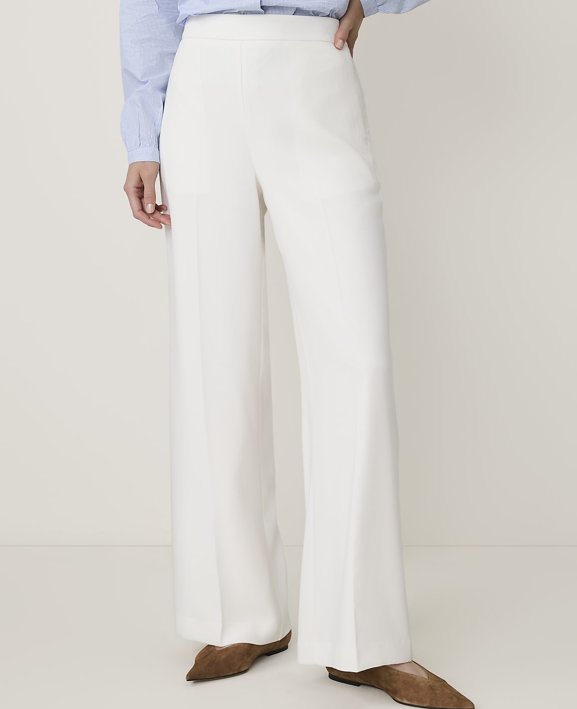 A person wearing white wide-leg pants and brown flats stands against a plain white background.