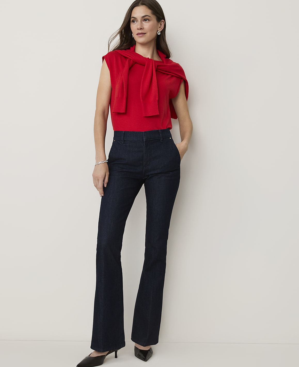 A woman in a red top and dark flared jeans stands against a plain white background.