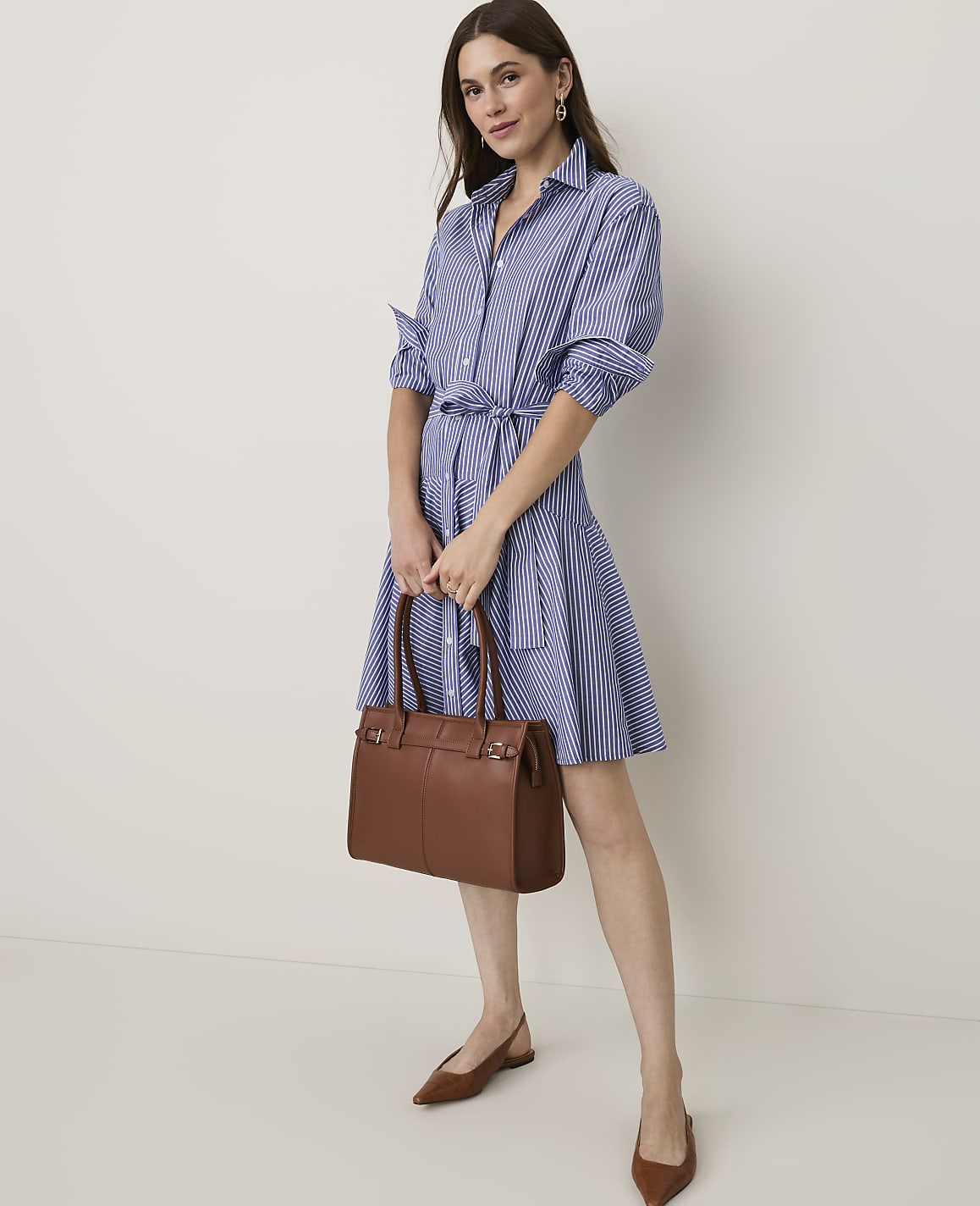 A woman in a blue and white striped shirt dress and brown flats holds a brown leather handbag in front of a plain white background.