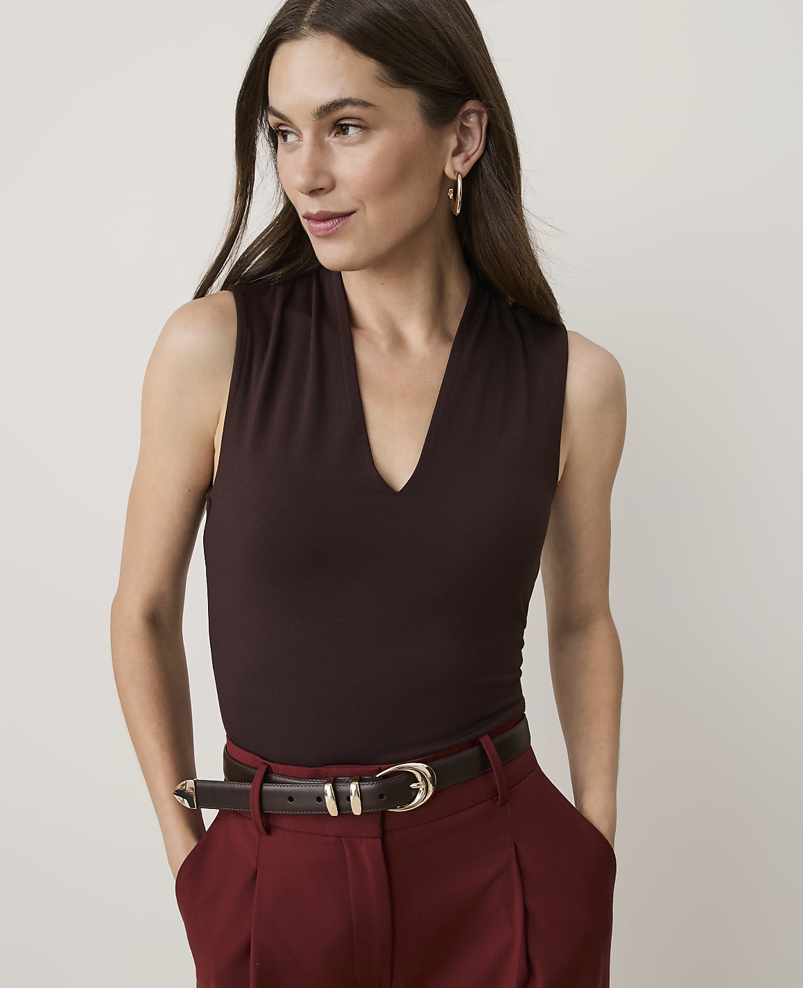A woman in a dark brown V-neck top and maroon pants stands against a plain background.
