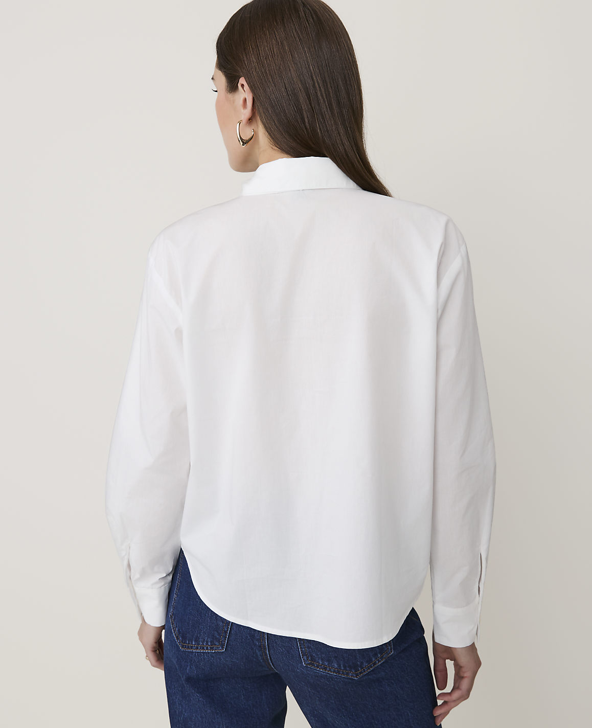 A woman in a white button-down shirt and blue jeans stands against a plain white background.