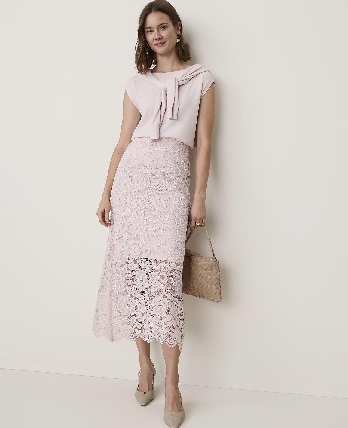 A woman in a light pink lace skirt and matching top stands against a plain, light-colored wall, holding a woven handbag.