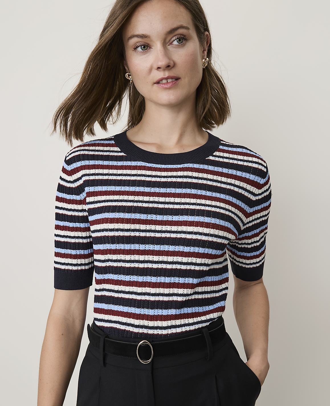 A woman in a striped sweater and black pants stands against a plain, light background.