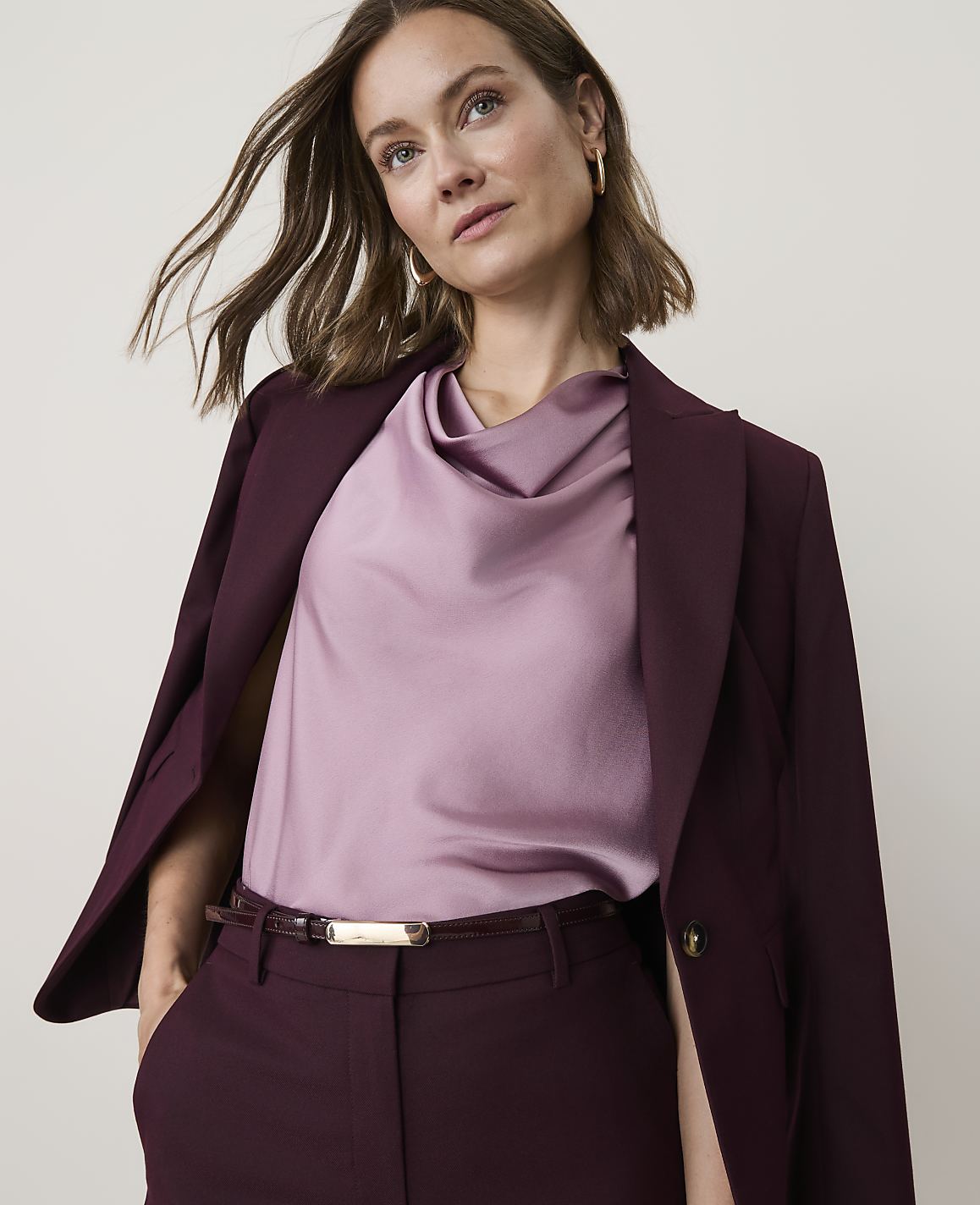 A woman in a burgundy suit and a lilac cowl-neck top stands against a plain, light background.