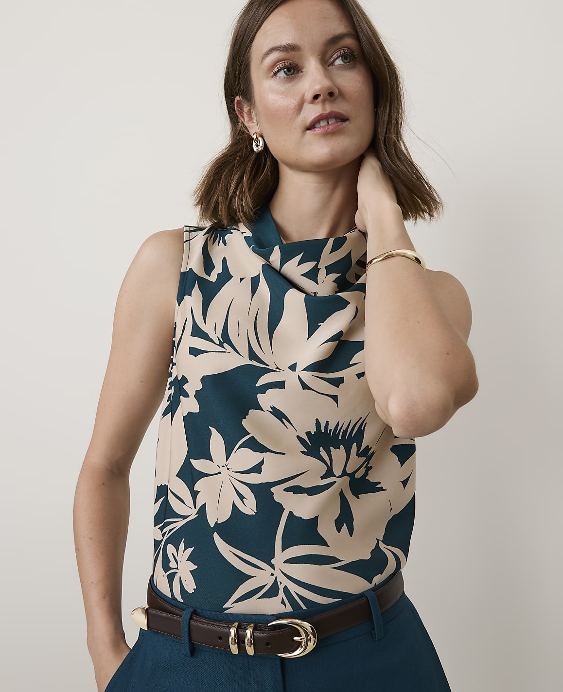 A woman in a floral top and dark pants stands against a plain, light background.