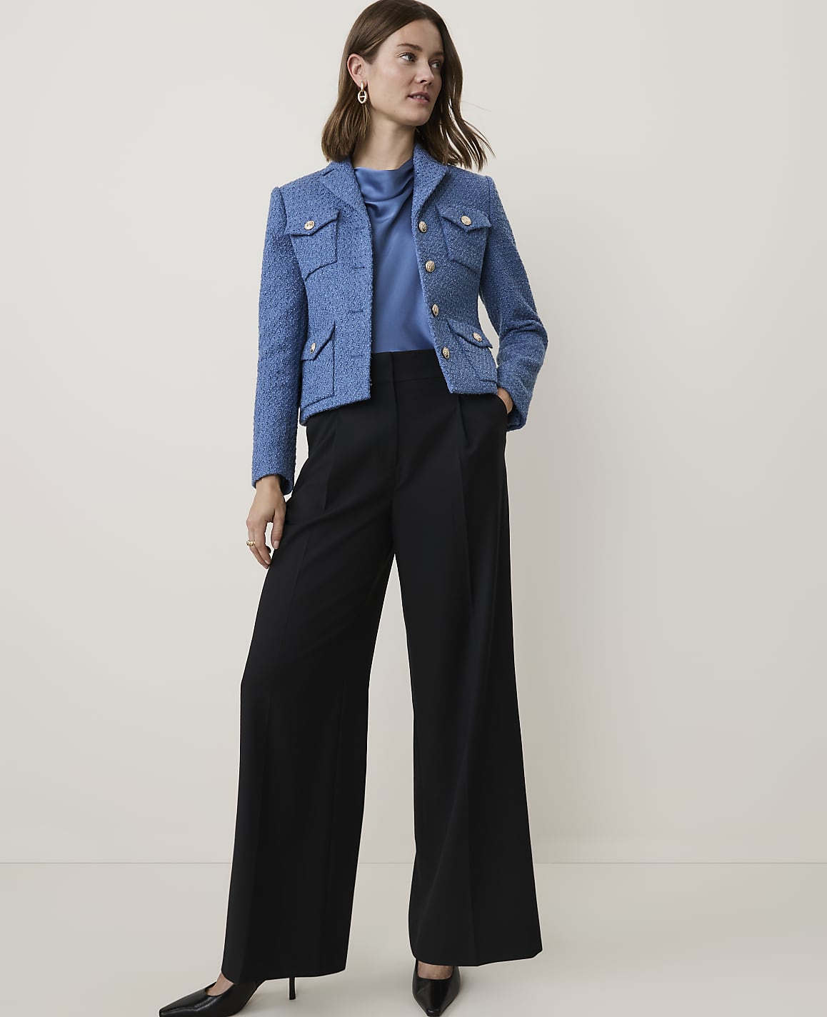 A woman in a blue tweed jacket and black wide-leg pants stands against a plain, light-colored background.