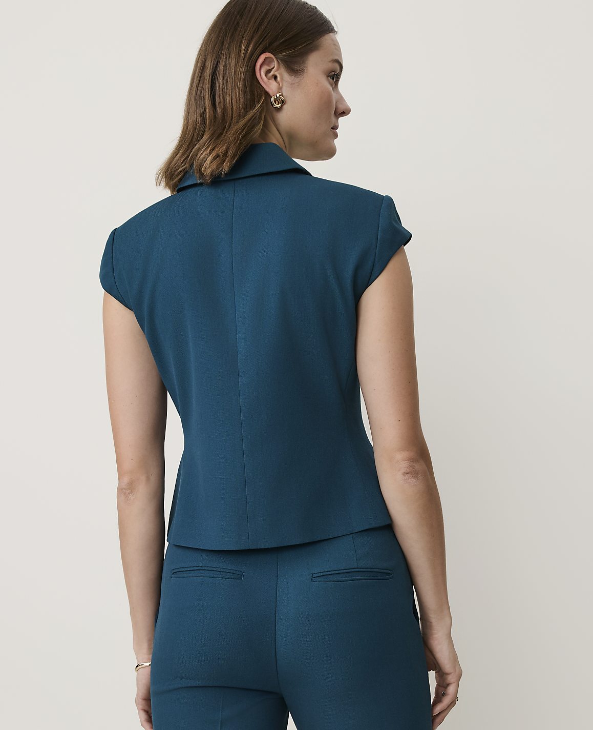 A woman in a teal blazer and pants stands against a plain, light background.