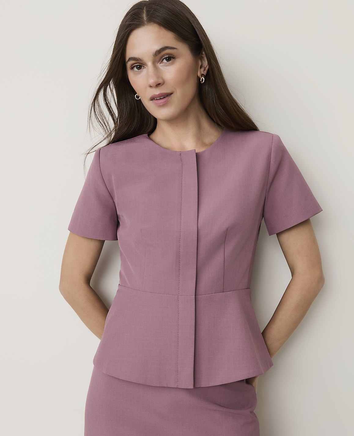 A woman in a mauve peplum jacket and skirt stands against a plain, light-colored background.