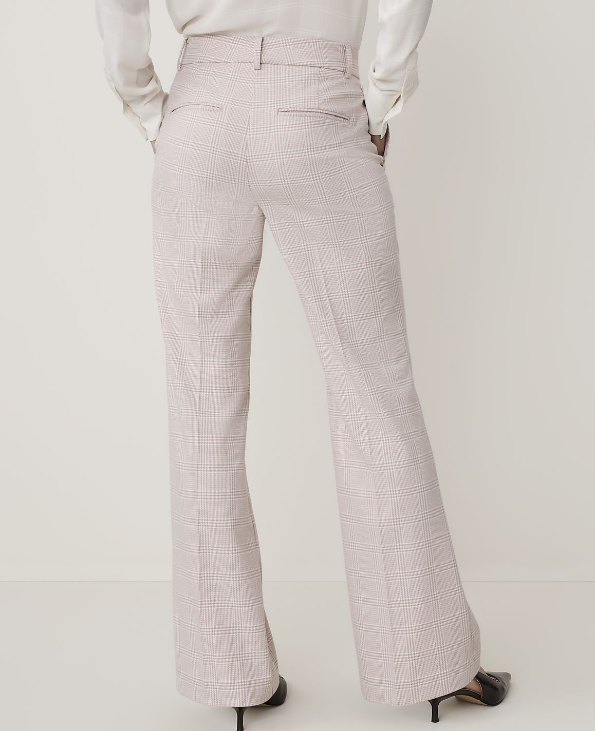 A person wearing light pink plaid flared trousers and a white blouse stands against a plain white background.