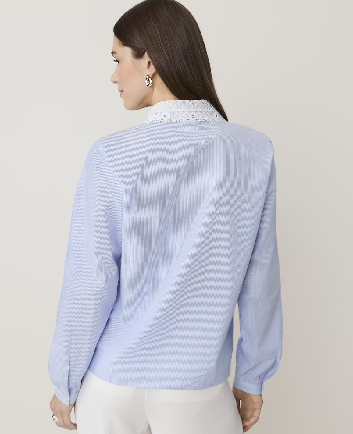 A woman in a light blue pinstripe blouse with lace trim stands against a plain, light background.