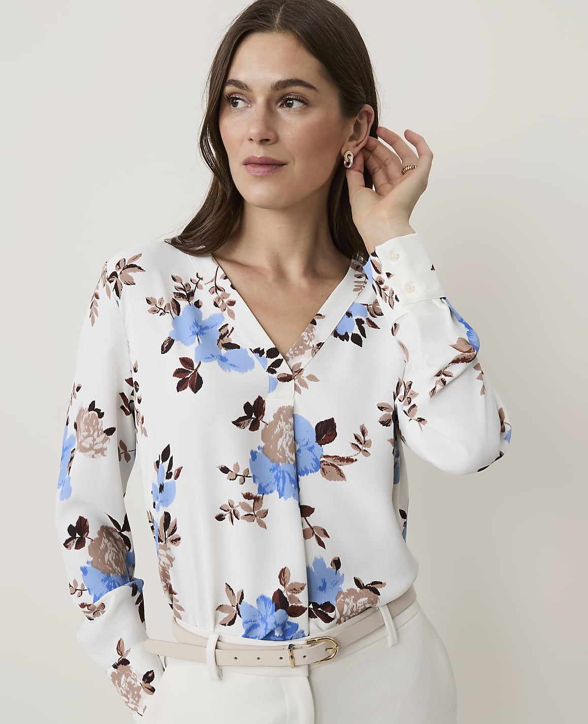 A woman in a floral blouse and cream pants stands against a plain white background.