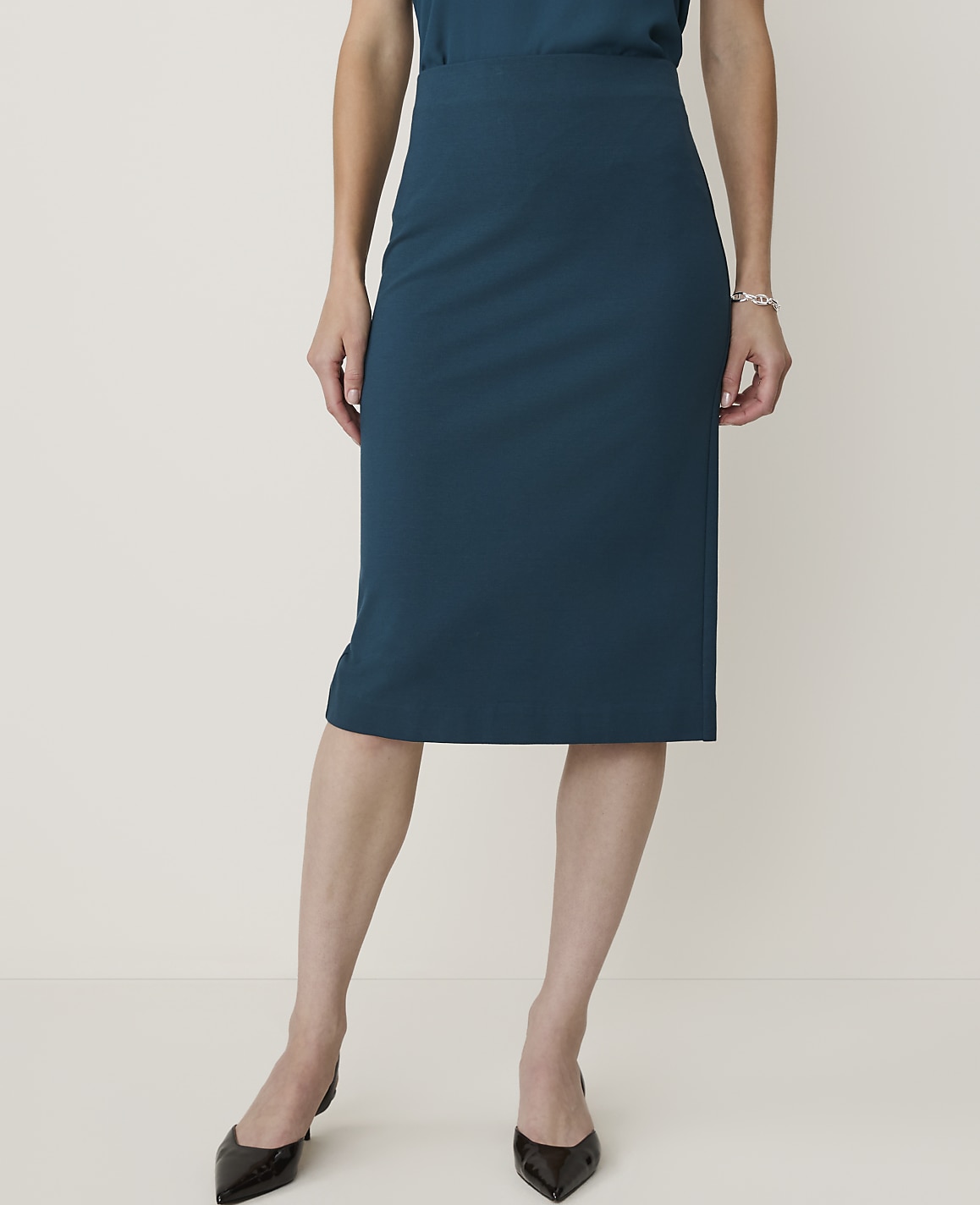 A woman in a teal pencil skirt and black pointed-toe heels stands against a plain, light background.
