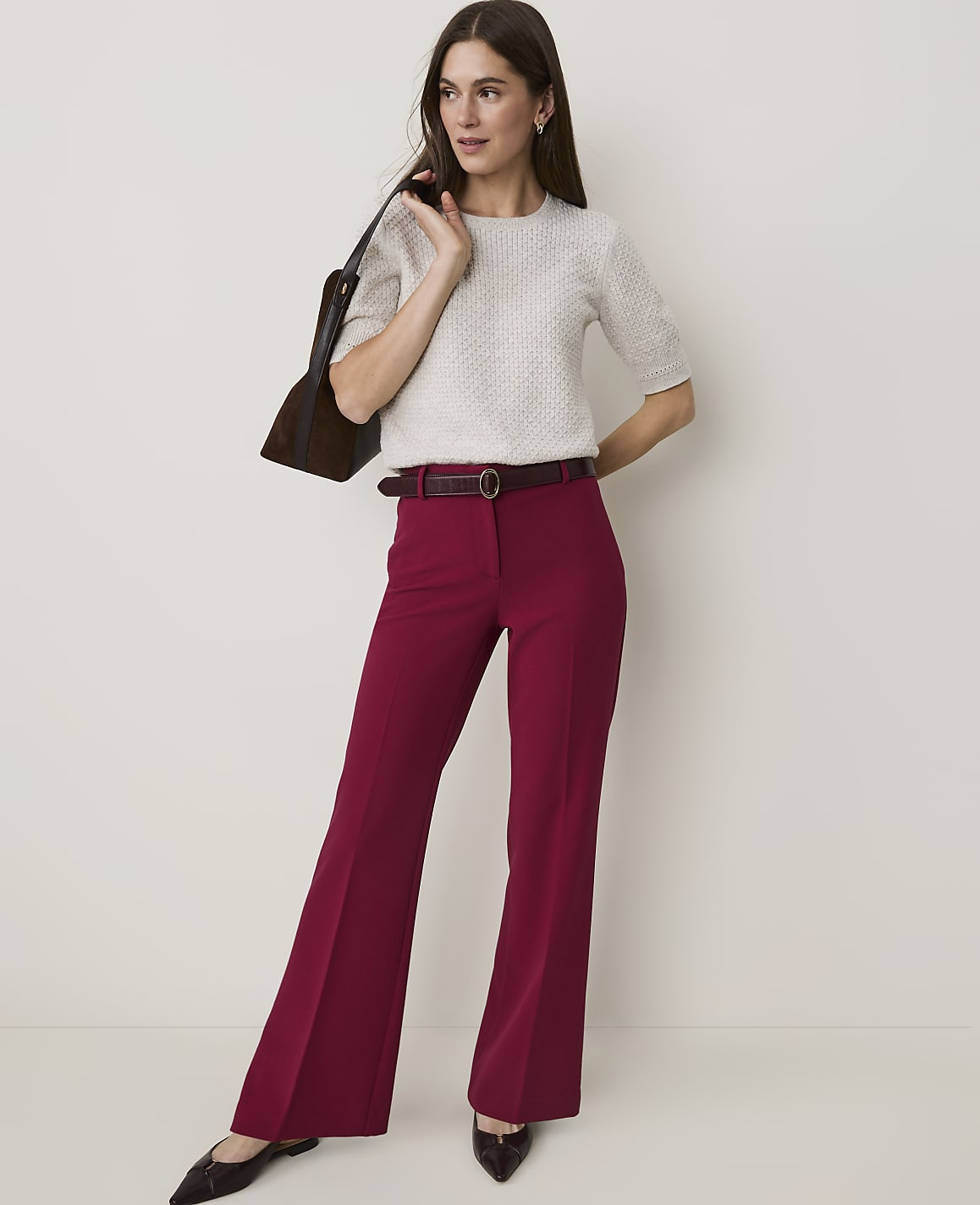 A woman in a cream sweater and burgundy flared pants stands against a plain, light background, carrying a dark brown bag.