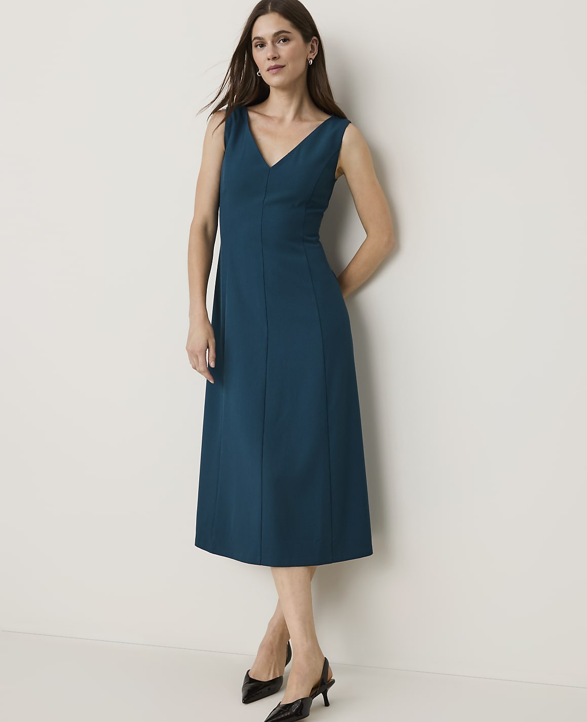 A woman in a teal V-neck dress and black heels stands against a plain, light-colored wall.